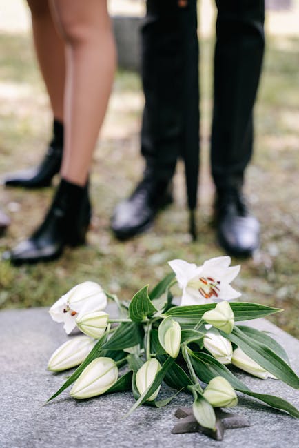 A bouquet of white lilies with large, open blooms displaying prominent stamens, surrounded by closed lily buds and dark green, elongated leaves, arranged on the ground. The flowers have smooth, velvet