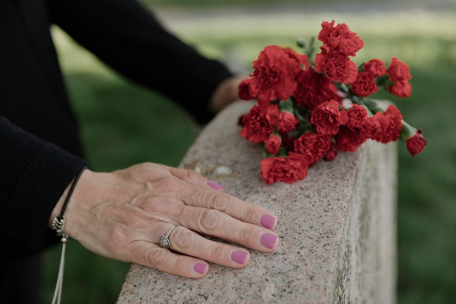 A hand with pink-painted nails and a silver ring rests on a stone surface next to a bouquet of red carnations. The carnations are freshly cut, with ruffled petals and green stems, arranged simply with