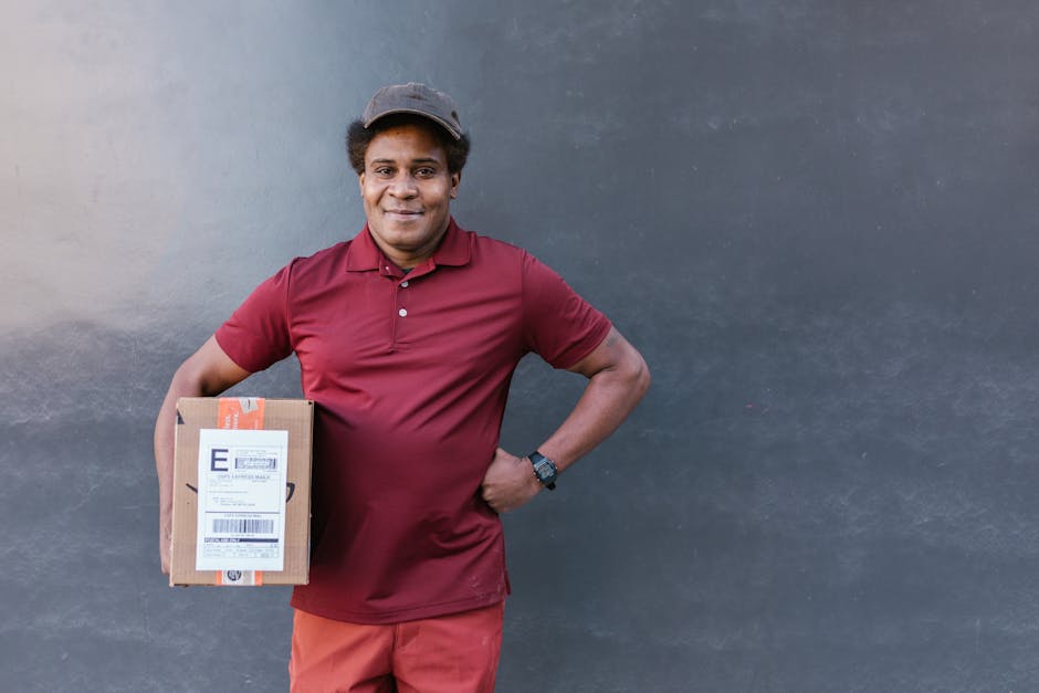 A smiling man with medium brown skin and short dark hair is standing outdoors against a plain, dark gray wall. He is wearing a maroon polo shirt, matching shorts, and a dark baseball cap. He holds a m