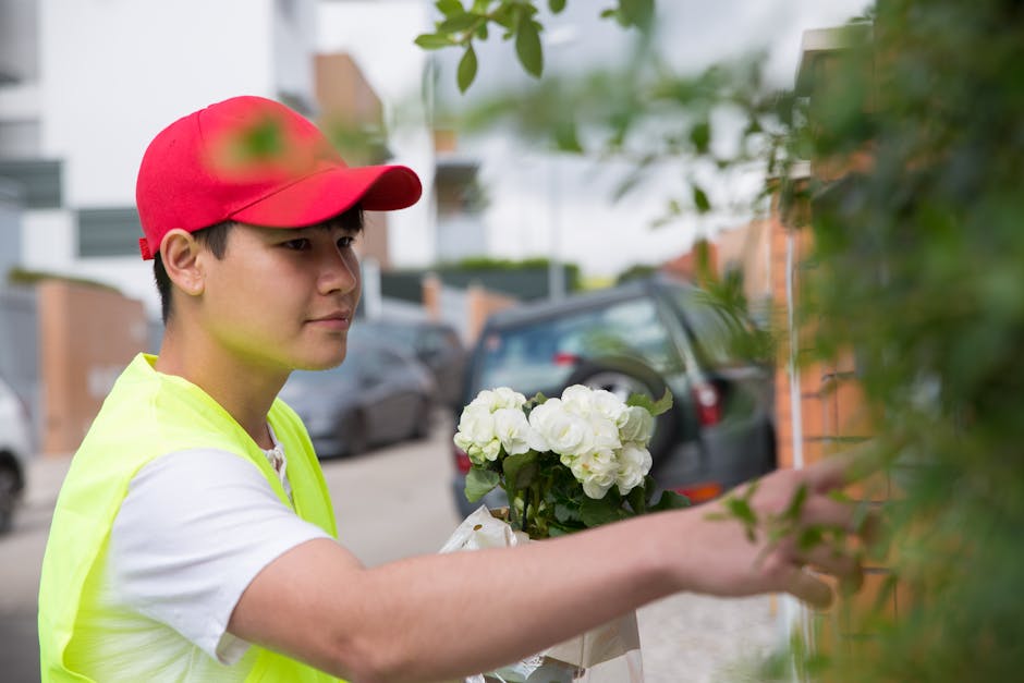 A young male florist wearing a red cap and a bright yellow high-visibility vest arranges a bouquet of fresh white roses and hydrangeas outdoors near a brick wall and parked cars. The bouquet is wrappe