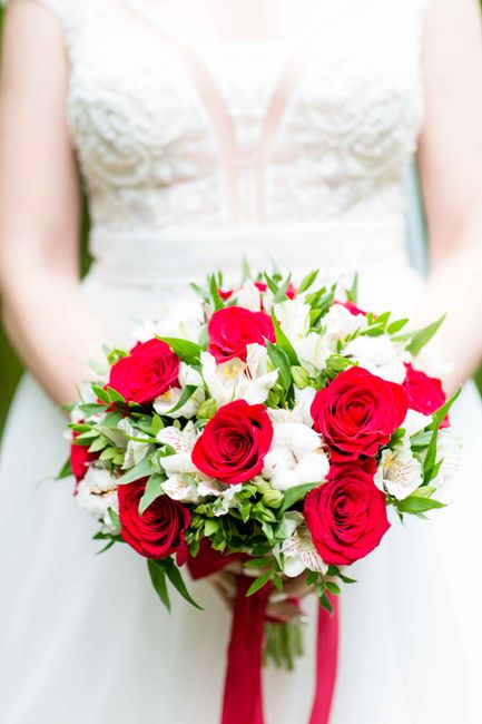 A bridal bouquet featuring vibrant red roses, white alstroemeria, and green foliage, arranged in a rounded, lush style. The flowers are fresh, with smooth petals and sturdy stems, held together with a
