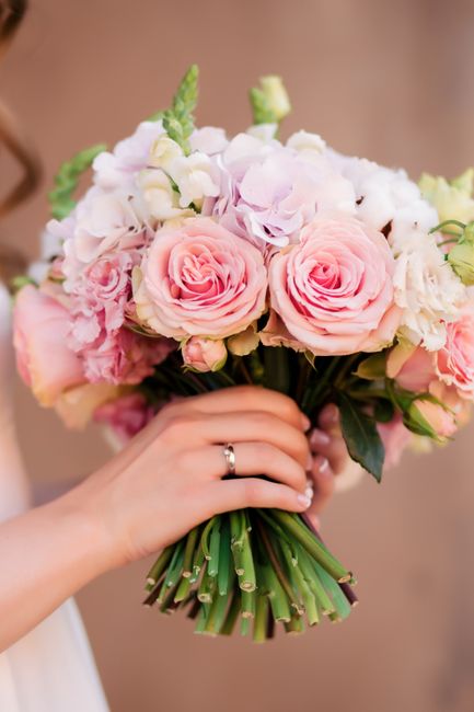 A person holding a bridal bouquet of fresh flowers featuring large, soft pink roses and pale lavender and white hydrangeas, with green foliage accents. The bouquet is tightly arranged with the stems g