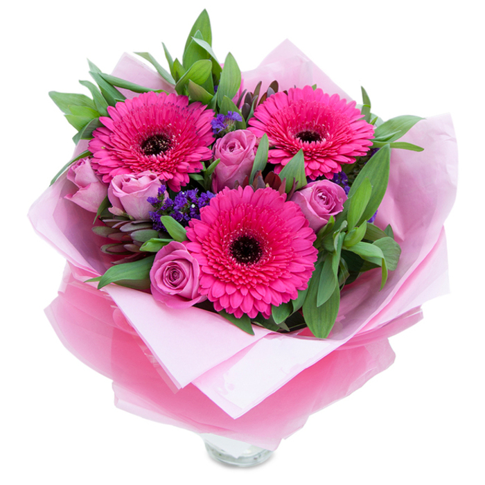 I see a bouquet that balances playful vibrancy with gentle grace, the sort of arrangement a local florist might tie up before a morning run to the High Street market. Dominating the scene are three vivid fuchsia Gerbera daisies, their radial petals opening like small suns around dark, almost black centres that add a dramatic pocket of contrast. Surrounding them are soft lavender-pink roses in early bloom, their tightly curled petals offering a velvety texture and a romantic softness that calms the composition. Sprays of tiny purple-blue statice tuck into the spaces between blooms, lending an airy, textural glitter that recalls wildflower edges near Langleybury fields. The greenery - broad salal-like leaves with the occasional rounded eucalyptus stem - frames the bouquet with a fresh, dewy green that enhances the pink palette and suggests careful selection for seasonal vibrancy. The wrapping is part of the craft: an inner layer of muted blush tissue paper provides a soft cradle, while an outer sheath of slightly crinkled, translucent fuchsia-pink cellophane catches the light and ties the whole presentation together with a subtle sheen. The florist's folds are neat and deliberate, and the clear glass vase rim visible at the bottom promises immediate display and water for lasting freshness. In soft daylight the petals seem tactile and plush, with roses lending a delicate perfume and the Gerberas offering a hint of green sweetness - an arrangement designed to celebrate small joys, from congratulations to a warm 'thinking of you' for someone in Abbots Langley.