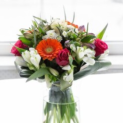A celebration of floral artistry and the verdant character of Abbots Langley, this stunning ensemble radiates warmth and grace. Creamy peach and vivid orange gerbera daisies set the tone, their rounded petals forming a soft halo that beckons the gaze. Beside them rest fuchsia roses, deep in hue and luxuriously dense, adding both brightness and sophistication to the overall look. White alstroemeria and dainty spray roses contribute layers of gentle intricacy, their many-petaled blooms emerging like morning mist on a village green. Subtle hints of pastel pink and bright white filler flowers weave through the composition, enhancing its fullness and depth. The bouquet is gathered within a protective embrace of glossy aspidistra leaves, curved and wrapped with a florist's precision-a nod to the thoughtful craftsmanship found throughout local shops near Gallows Hill Lane. Every stem stands visible in the transparent glass vase, the pale light from a nearby window amplifying the colours without overpowering their delicate nature. There is an almost palpable freshness to this arrangement, evoking early spring mornings and the first blossoms along Abbots Langley's quiet lanes. This bouquet, imbued with optimism and gentle care, feels perfect for marking new beginnings or expressing heartfelt thanks within the local community. Its harmonious structure, colour, and texture come together as an ideal centrepiece for gatherings, everyday joy, or simply sharing a piece of lovingly arranged Abbots Langley beauty.